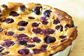 Un clafoutis aux cerises, dessert traditionnel français, présenté dans un moule à tarte blanc, avec des cerises noires réparties sur une pâte dorée et moelleuse, illustrant un classique gourmand de la pâtisserie maison.